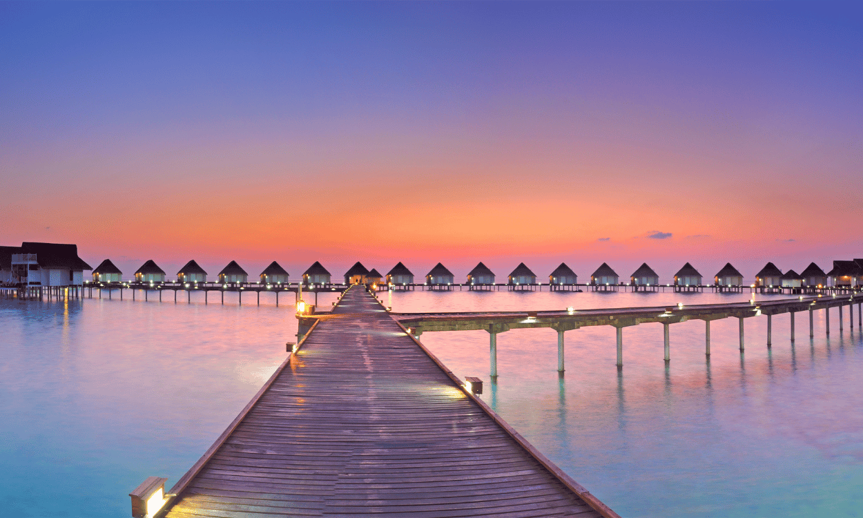 Holzsteg führt zu Wasserbungalows im Meer unter einem farbenprächtigen Sonnenuntergang. Lichter spiegeln sich im ruhigen Wasser. Holzsteg führt zu Wasserbungalows im Meer unter einem farbenprächtigen Sonnenuntergang. Lichter spiegeln sich im ruhigen Wasser.
