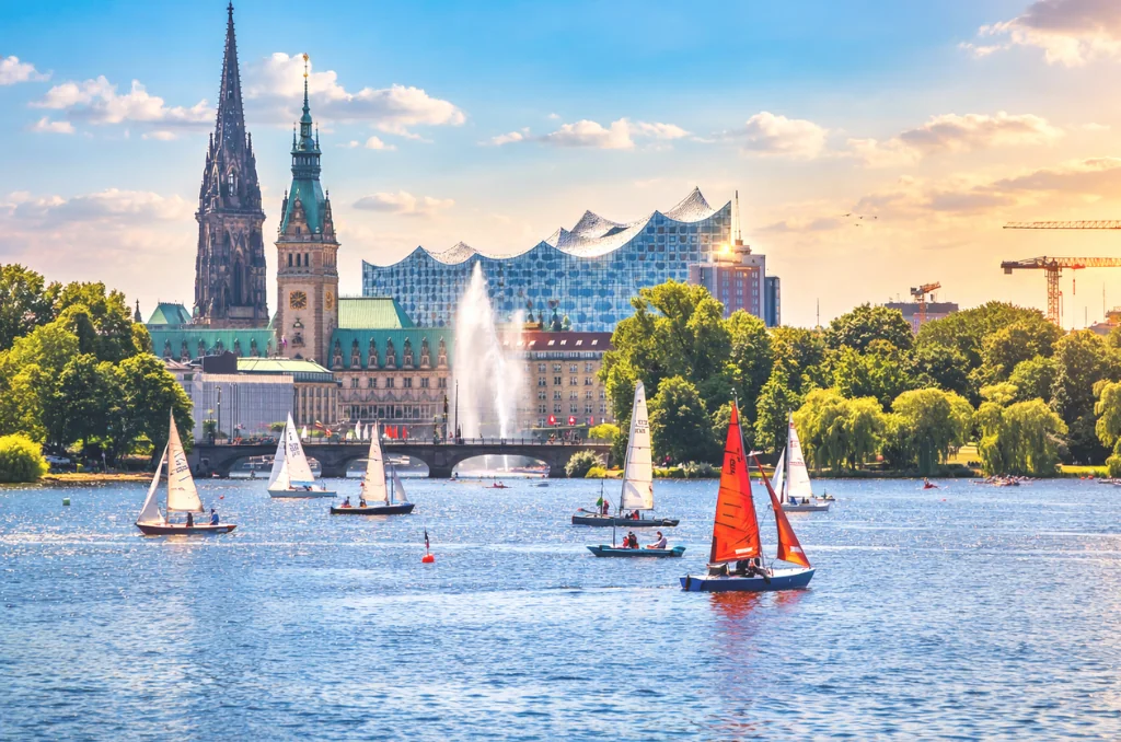 Ein sonniger Tag auf der Hamburger Außenalster mit Segelbooten und der berühmten Skyline im Hintergrund, inklusive Elbphilharmonie und Rathaus.